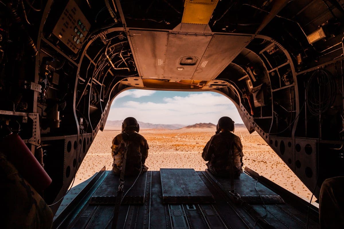 Two soldiers seated in the open rear of a military aircraft over desert