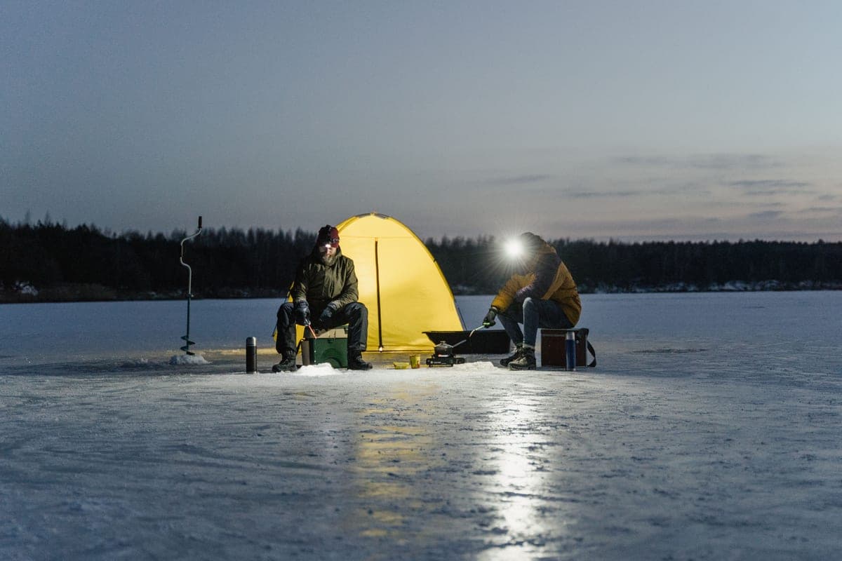 Two people beside a tent on a frozen lake at dusk, one wearing a headlamp
