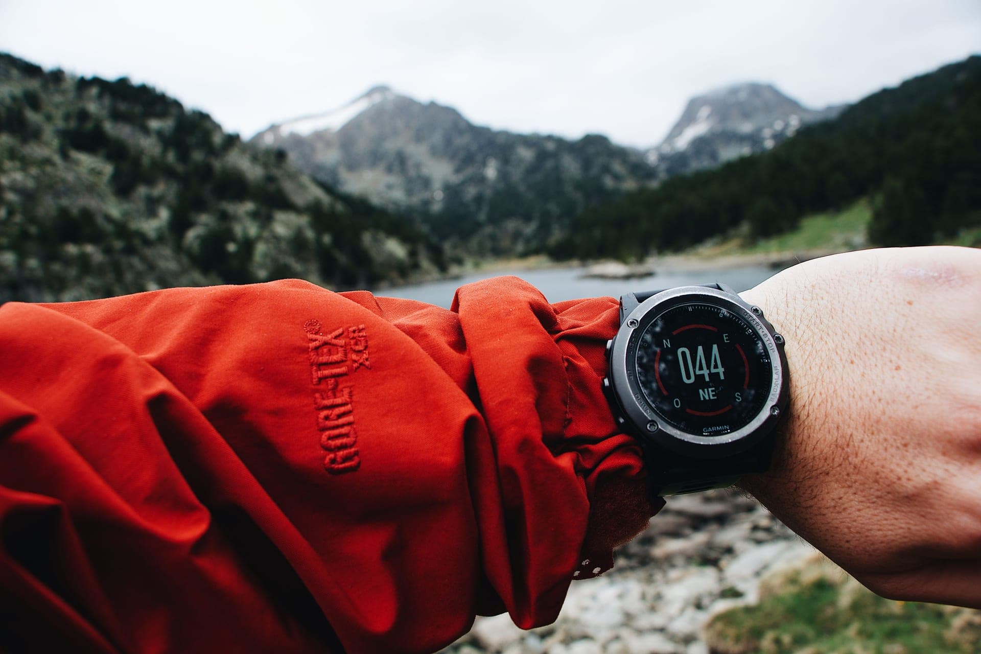 A GPS watch showing a compass bearing on a wrist in a red shell jacket, with mountains and a lake behind